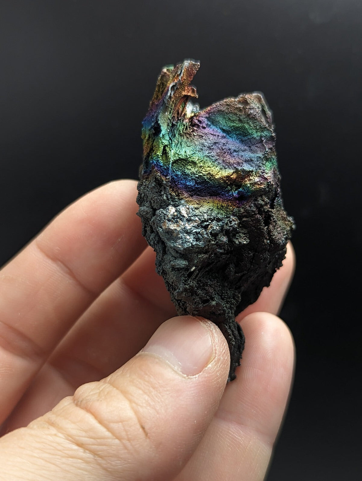 A hand holds a rough, irregularly shaped piece of iridescent mineral with vibrant rainbow hues reflecting off its surface against a dark background.