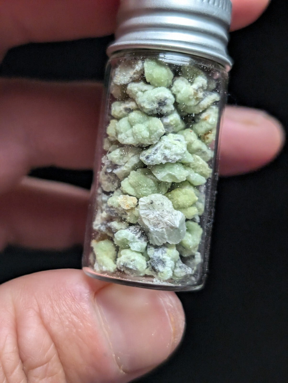 A clear glass vial with a silver metal cap contains small, irregularly shaped greenish-white crystalline pieces.