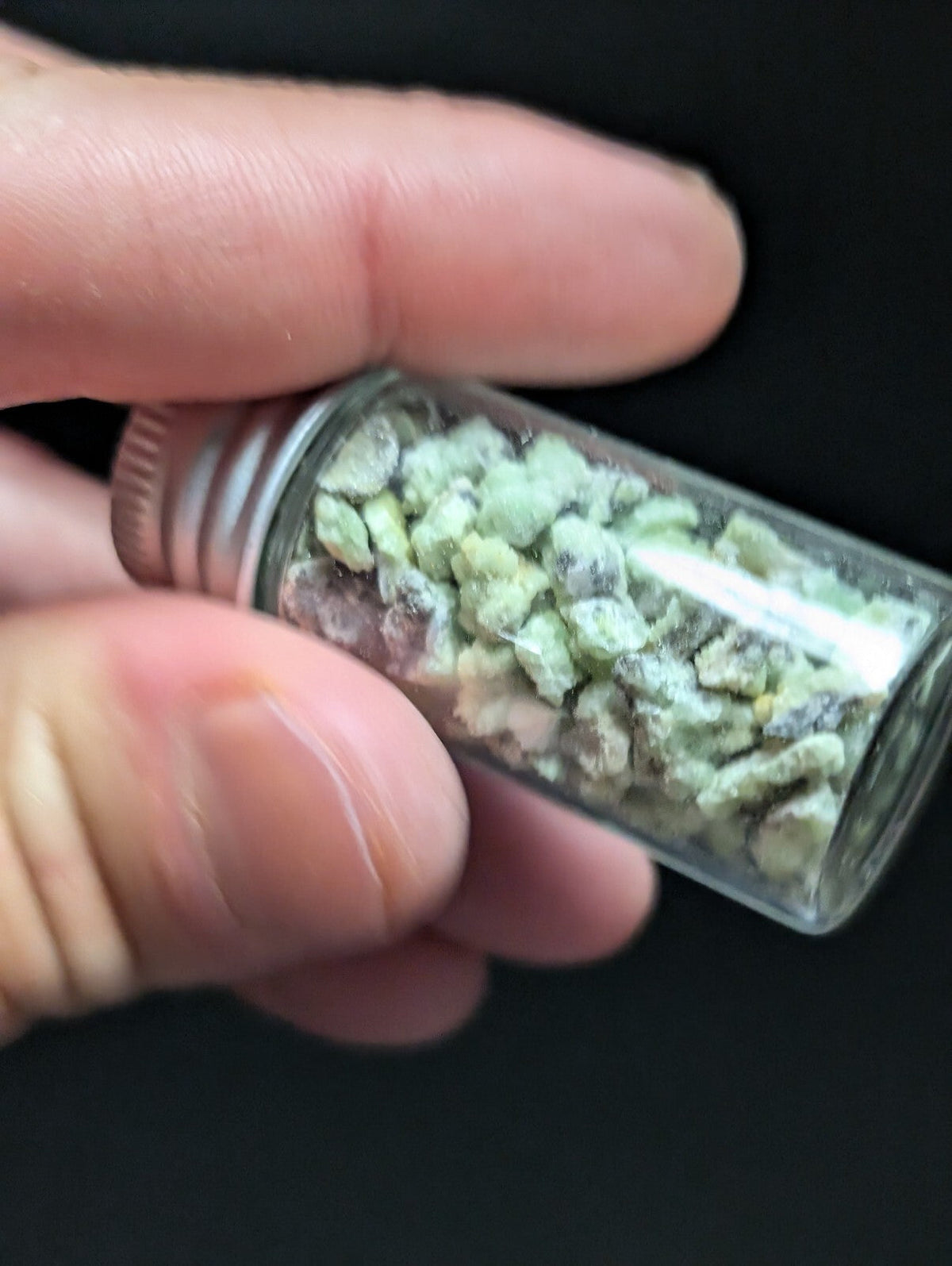 A clear glass vial with a silver metal cap, filled with small greenish-white crystalline fragments.