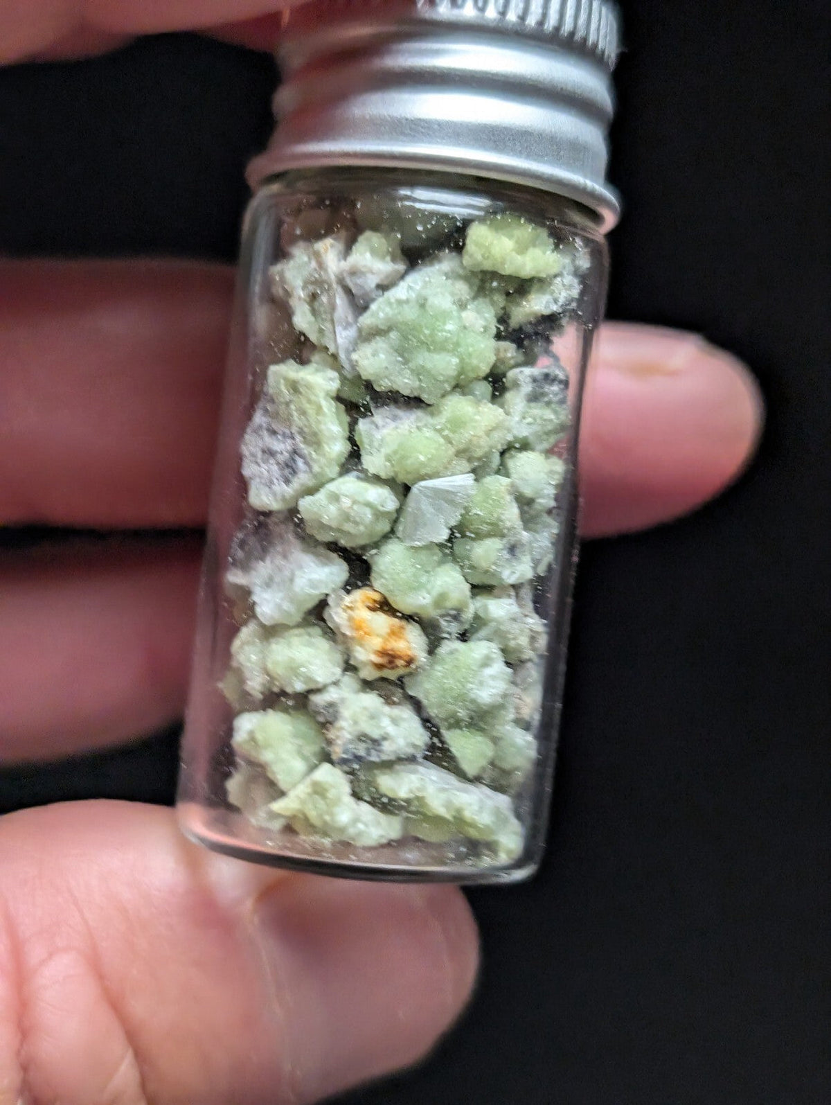 A clear glass vial with a silver metal screw-top lid contains small, irregularly shaped green mineral fragments.