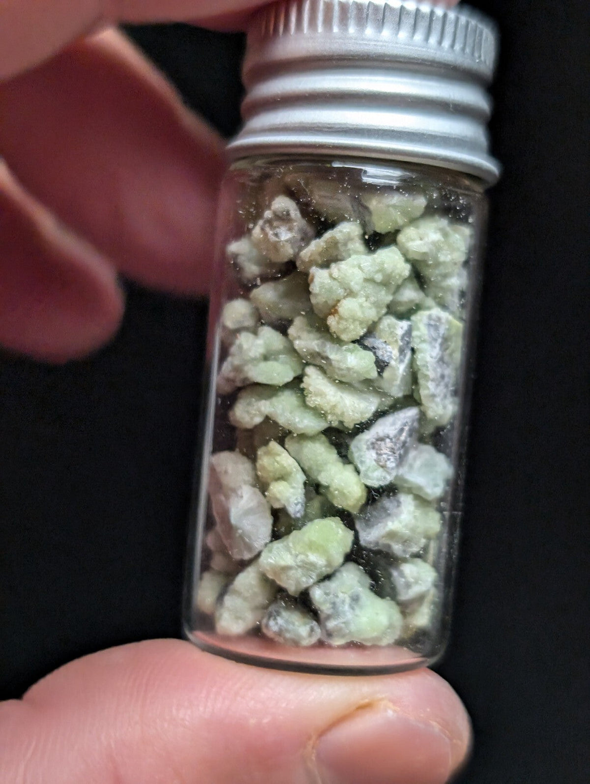 A clear glass vial with a silver metal screw-top lid contains irregularly shaped, pale greenish-white mineral fragments.