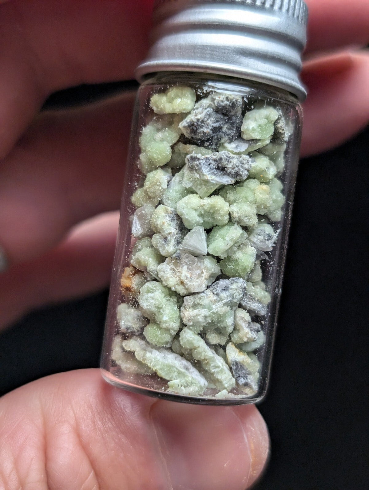 A clear glass vial with a silver metal screw-top lid contains small, irregularly shaped green and gray mineral fragments.