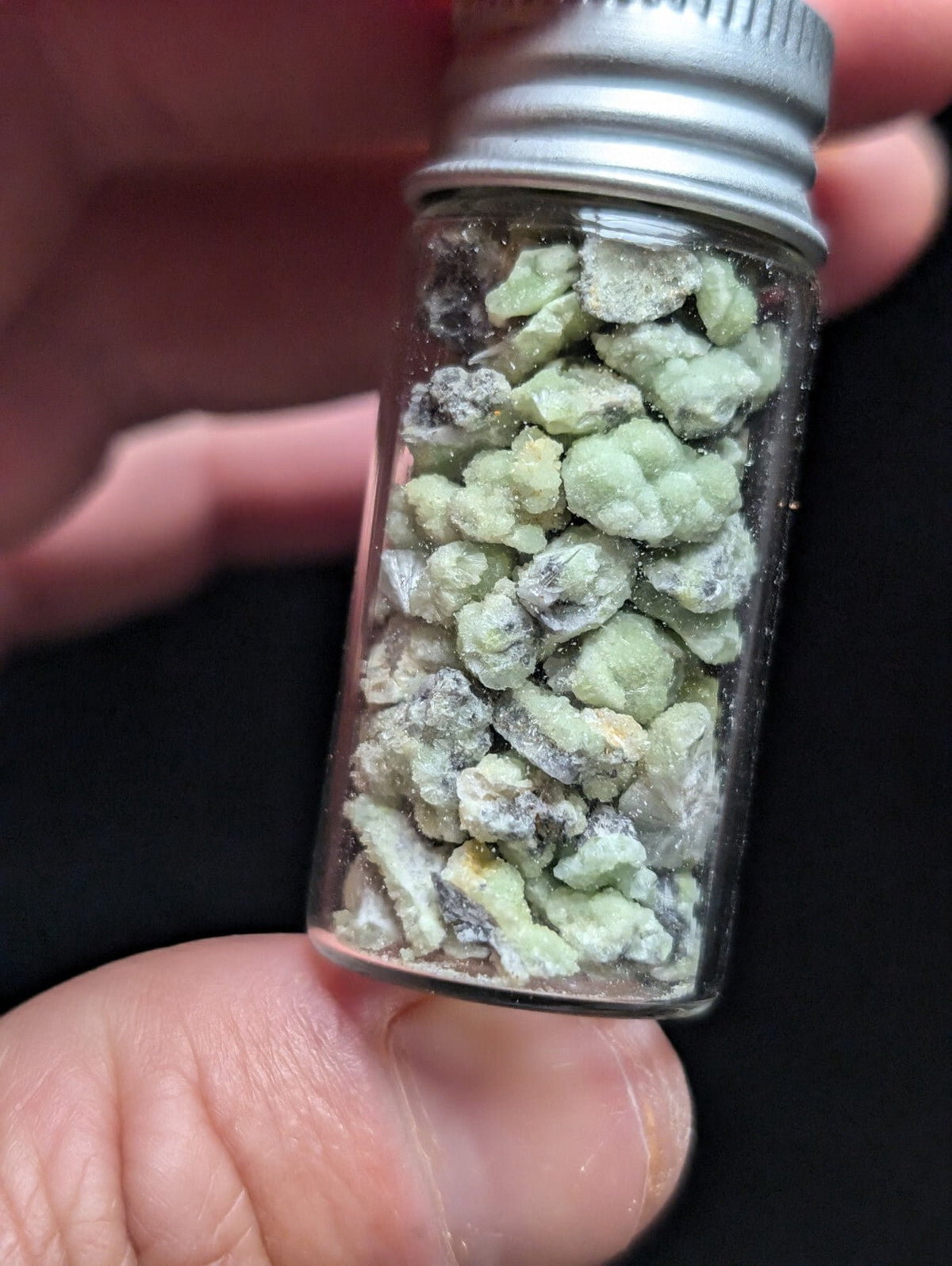A clear glass vial with a silver metal screw-top lid contains small, irregularly shaped greenish-white chunks that appear to be natural mineral specimens.