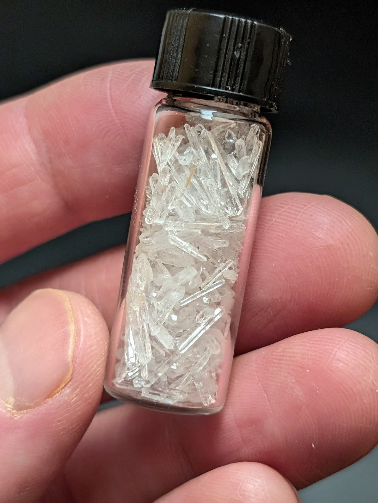 A clear glass vial with a black ribbed cap contains numerous transparent, crystalline shards.