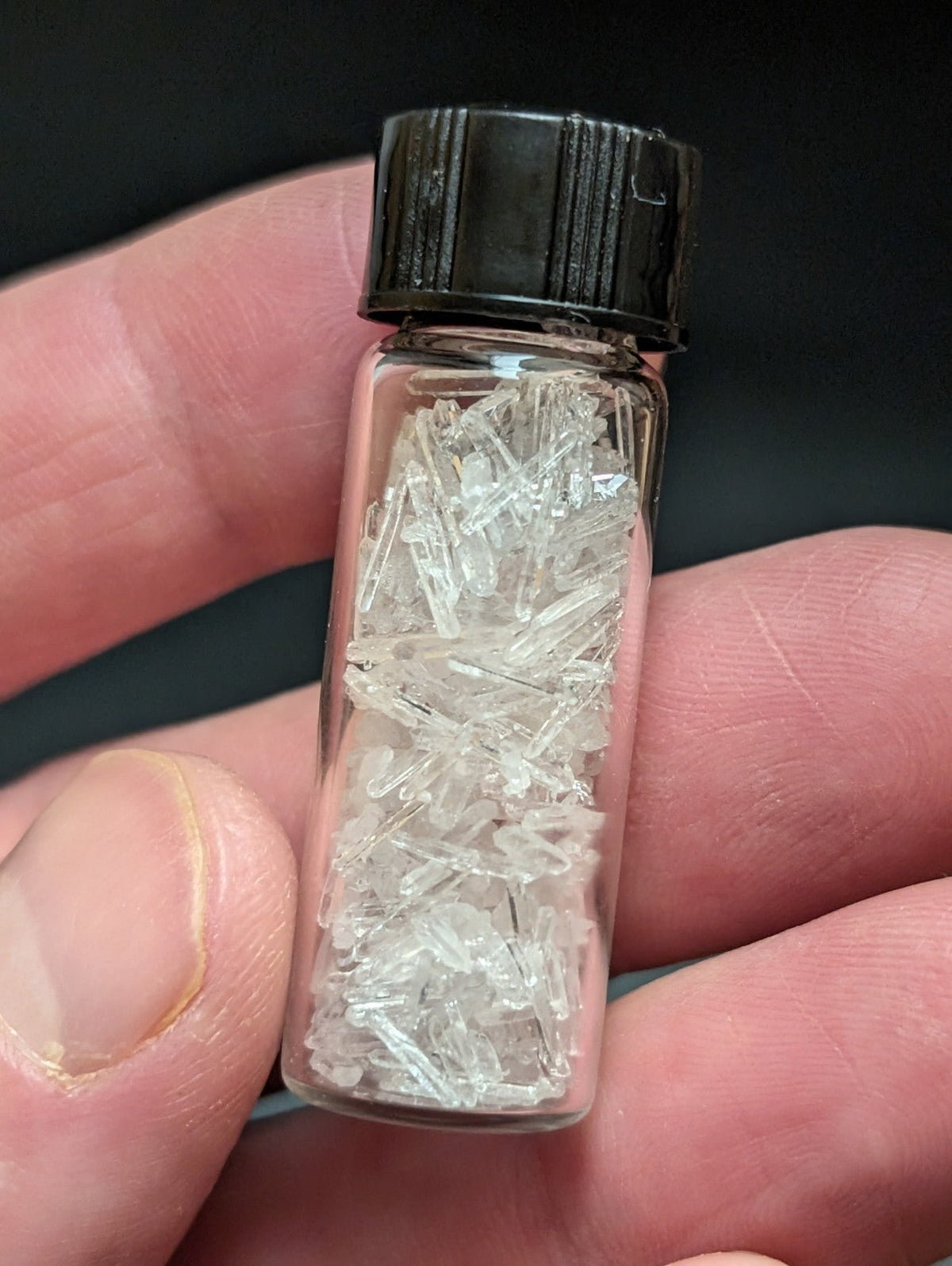 A clear glass vial with a black ribbed cap contains numerous transparent, crystalline fragments.