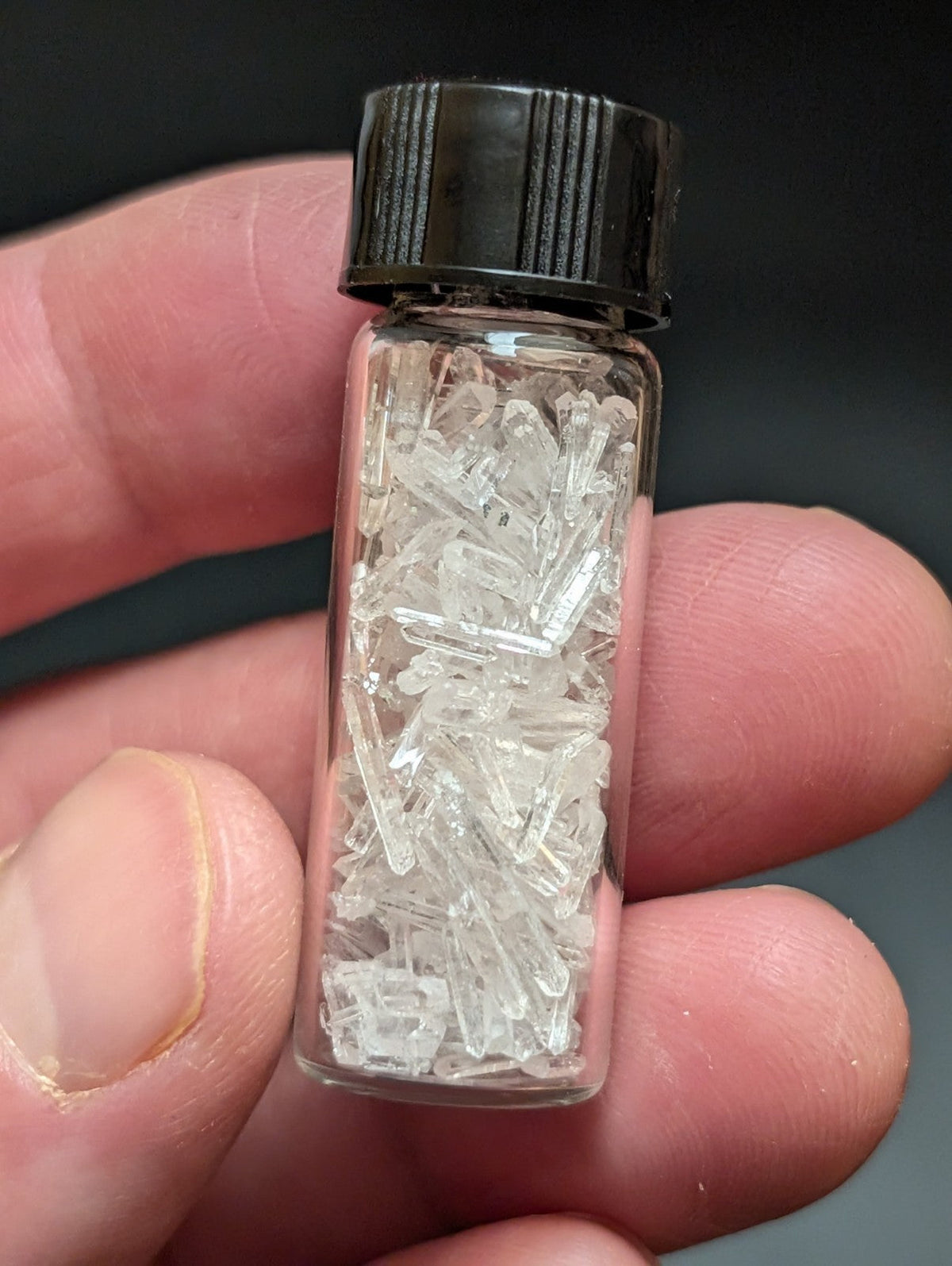 A clear glass vial with a black ribbed cap contains numerous transparent, crystalline fragments.