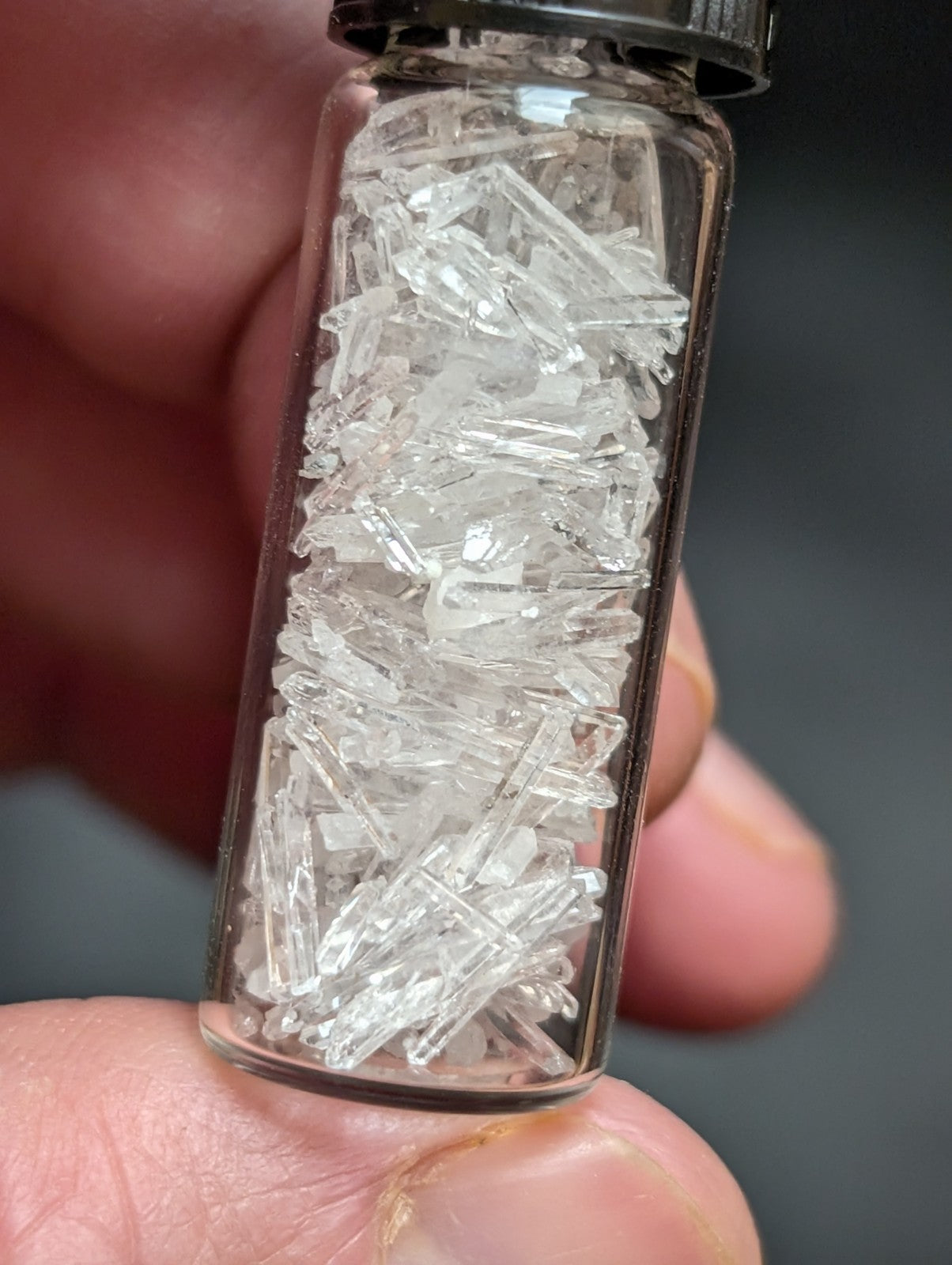A clear glass vial with a black screw-top lid, filled with transparent, crystalline fragments.