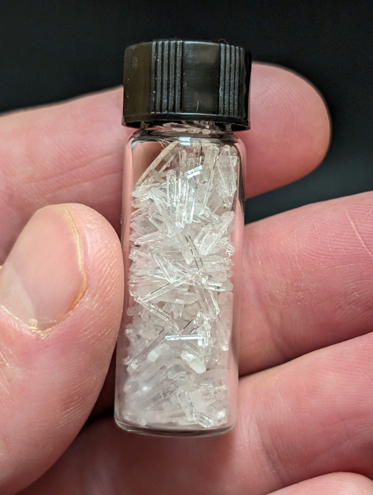 A clear glass vial with a black ribbed cap contains numerous transparent, crystalline fragments.