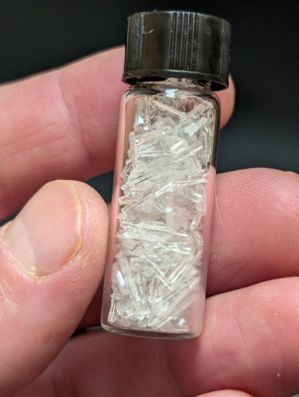 A clear glass vial with a black ribbed cap contains numerous transparent, crystalline fragments.