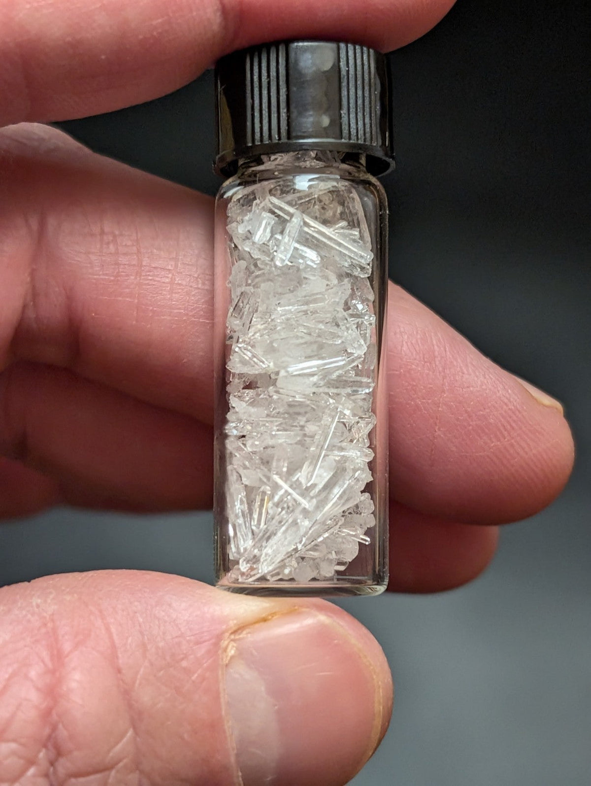A clear glass vial with a black ribbed cap, filled with transparent crystalline fragments.