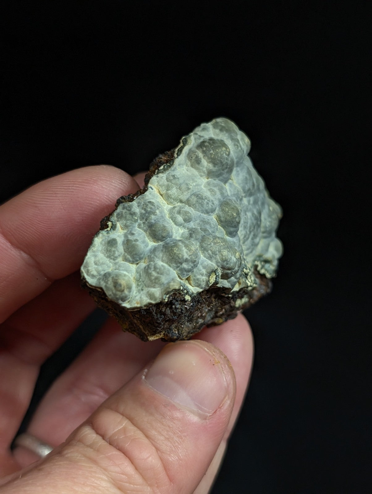 A hand holds a rough, irregularly shaped piece of greenish-gray mineral with a textured surface featuring rounded nodules.