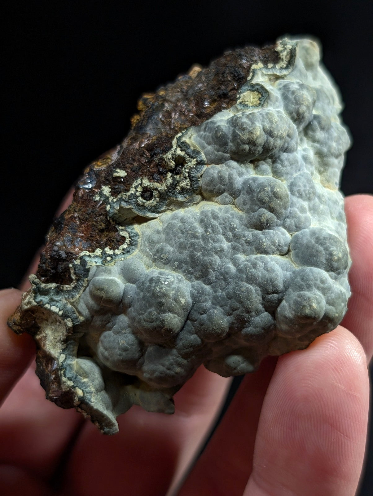 A textured, grayish-green mineral specimen with a bumpy surface and patches of darker brown lichen.
