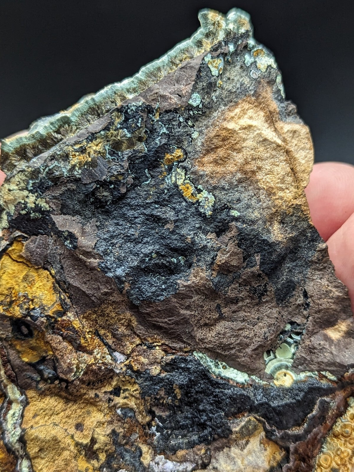 A rugged, irregularly shaped mineral specimen featuring a striking mix of dark black and earthy brown tones with vibrant patches of greenish-blue and golden-yellow crystalline formations.