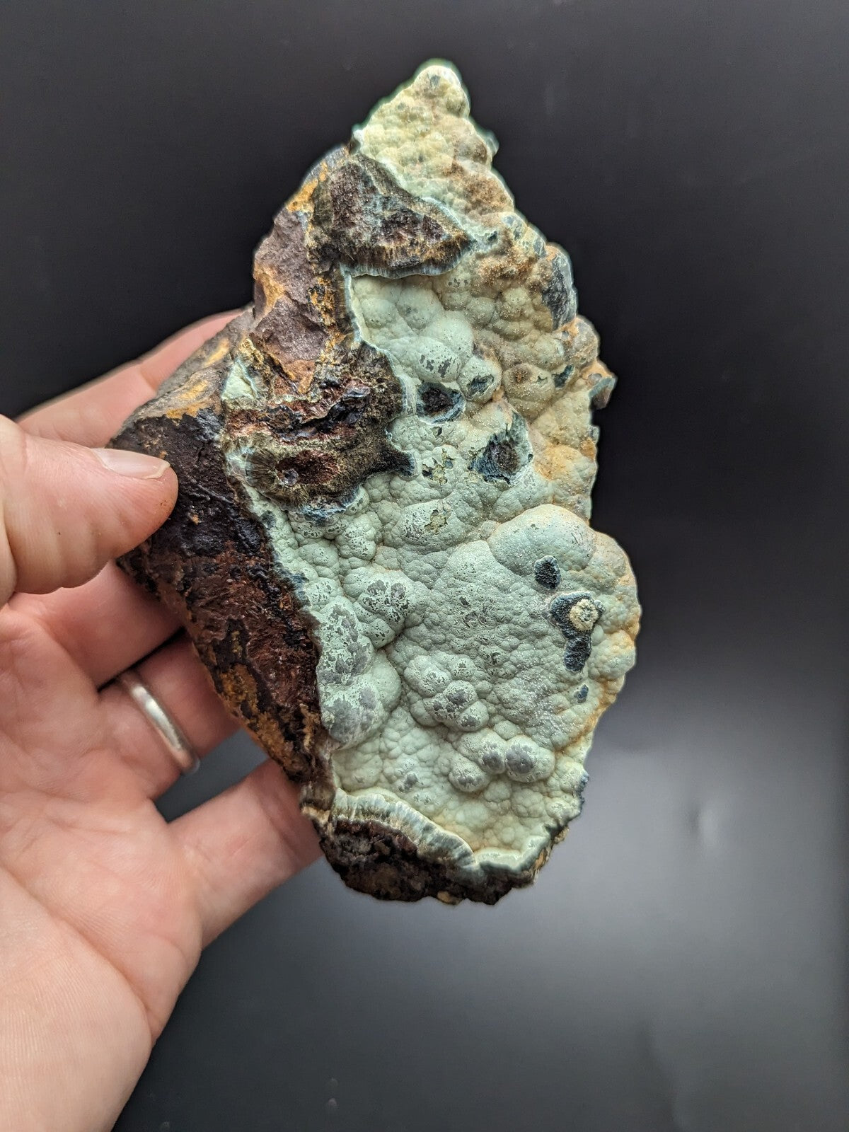 A hand holds a rugged, irregularly shaped piece of mineral with a textured surface featuring patches of pale greenish-blue lichen-like growths over darker brown and rust-colored rock.