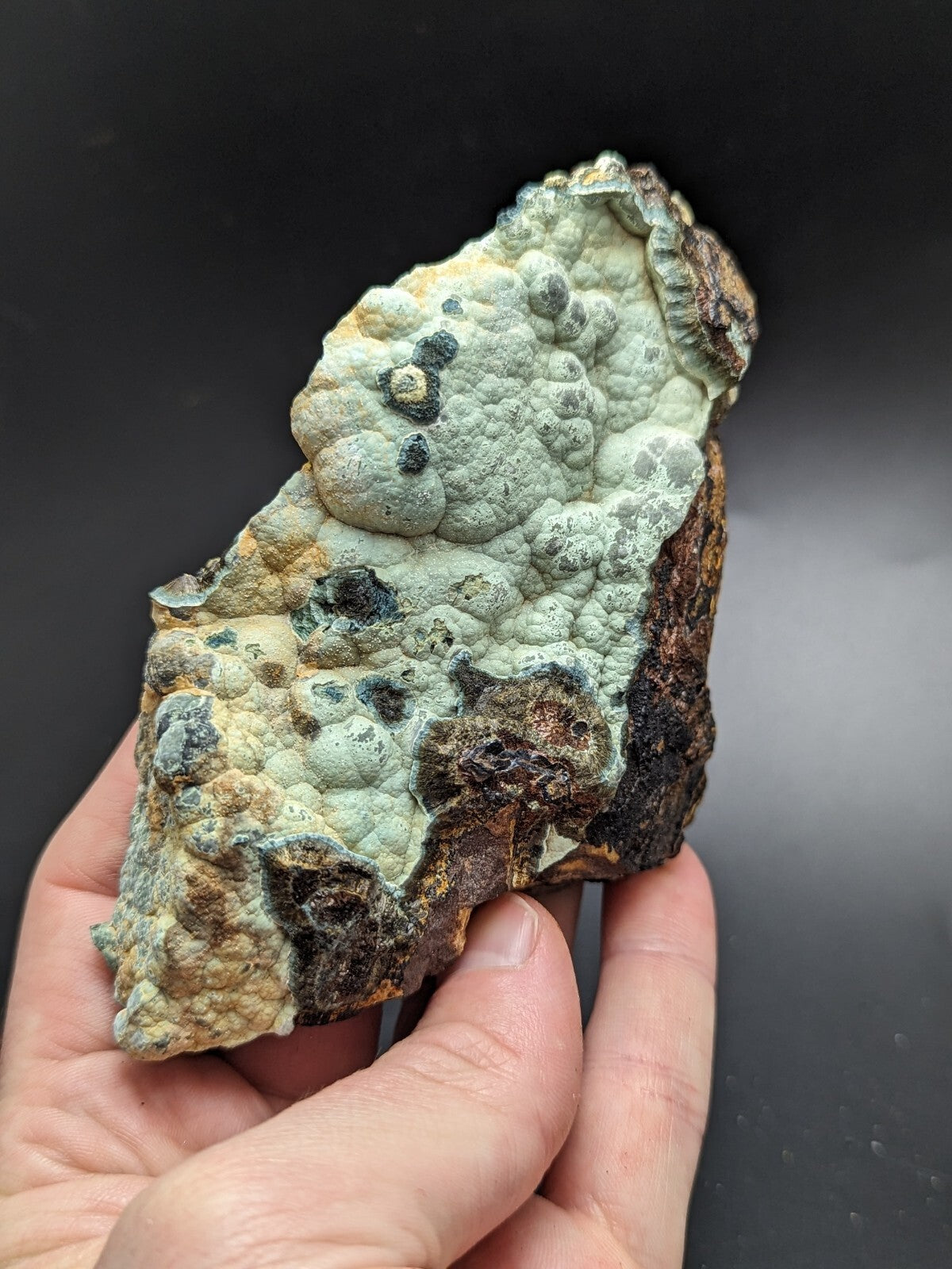 A textured, irregularly shaped mineral specimen with a pale greenish-blue surface and patches of dark brown and black, held in someone’s hand.