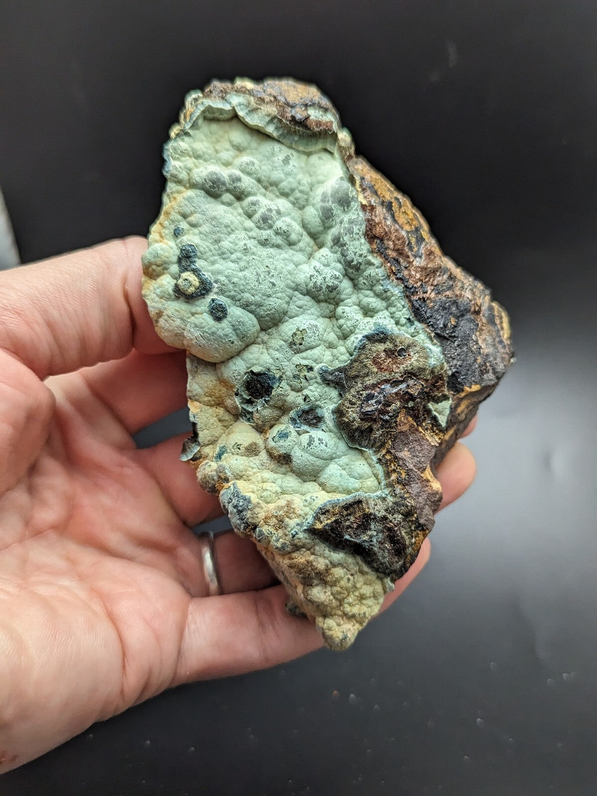 A hand holds a rough, irregularly shaped mineral specimen featuring patches of pale green and brownish-black with textured, bumpy surfaces.