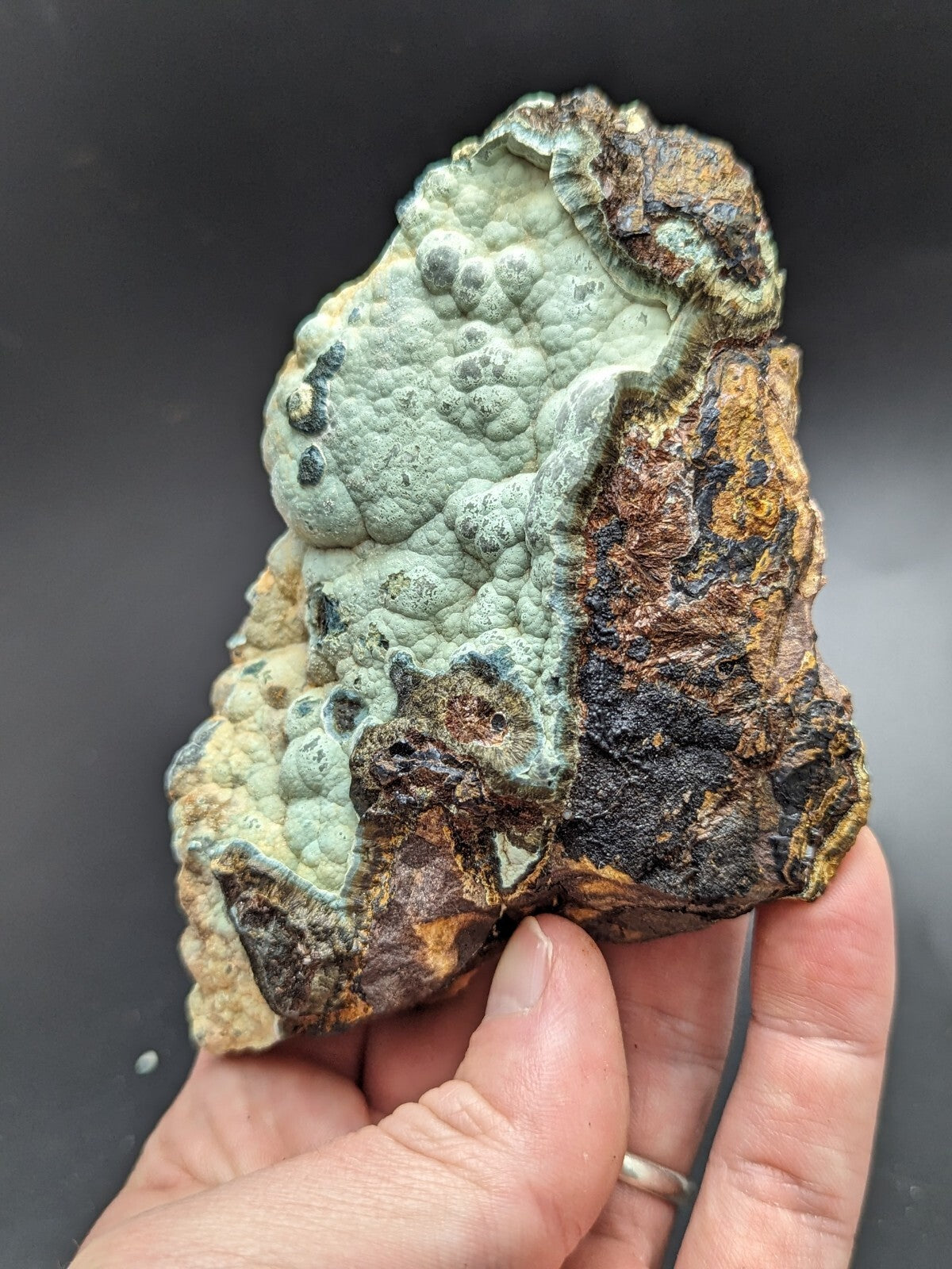 A hand holds a rugged, irregularly shaped mineral specimen featuring patches of pale greenish-blue lichen-like formations over dark brown and blackened rock.