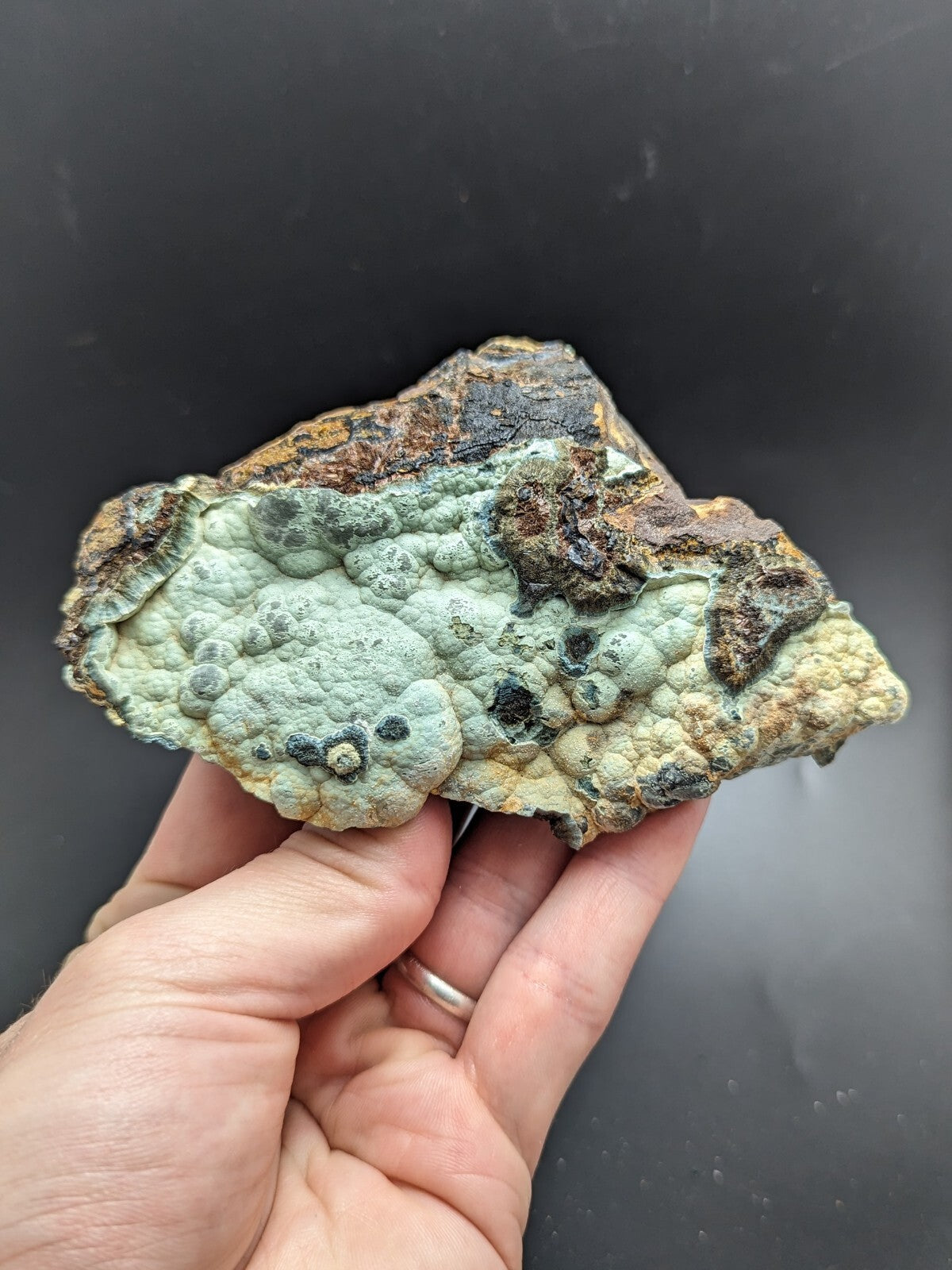 A hand holds a textured, irregularly shaped piece of mineral with patches of light green and pale yellow lichen-like growths over darker brown and black areas.