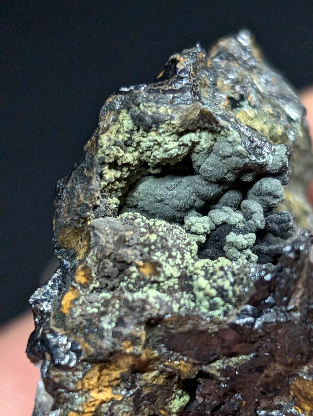A rugged, dark gray rock specimen with patches of vibrant green lichen and streaks of yellow mineral deposits.