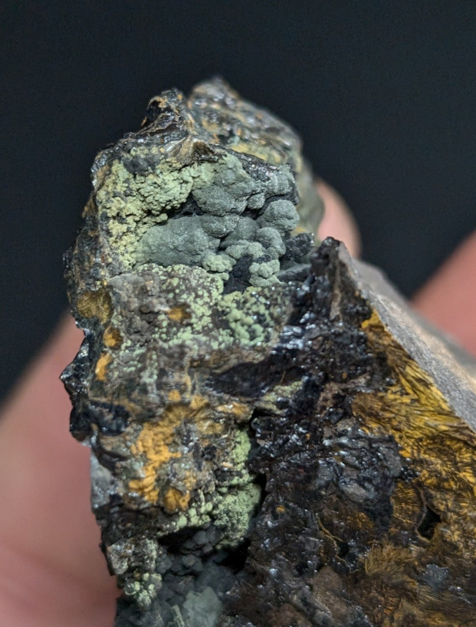 A rugged, irregularly shaped mineral specimen featuring patches of greenish lichen-like growths over a dark, textured base with streaks of yellow and black.