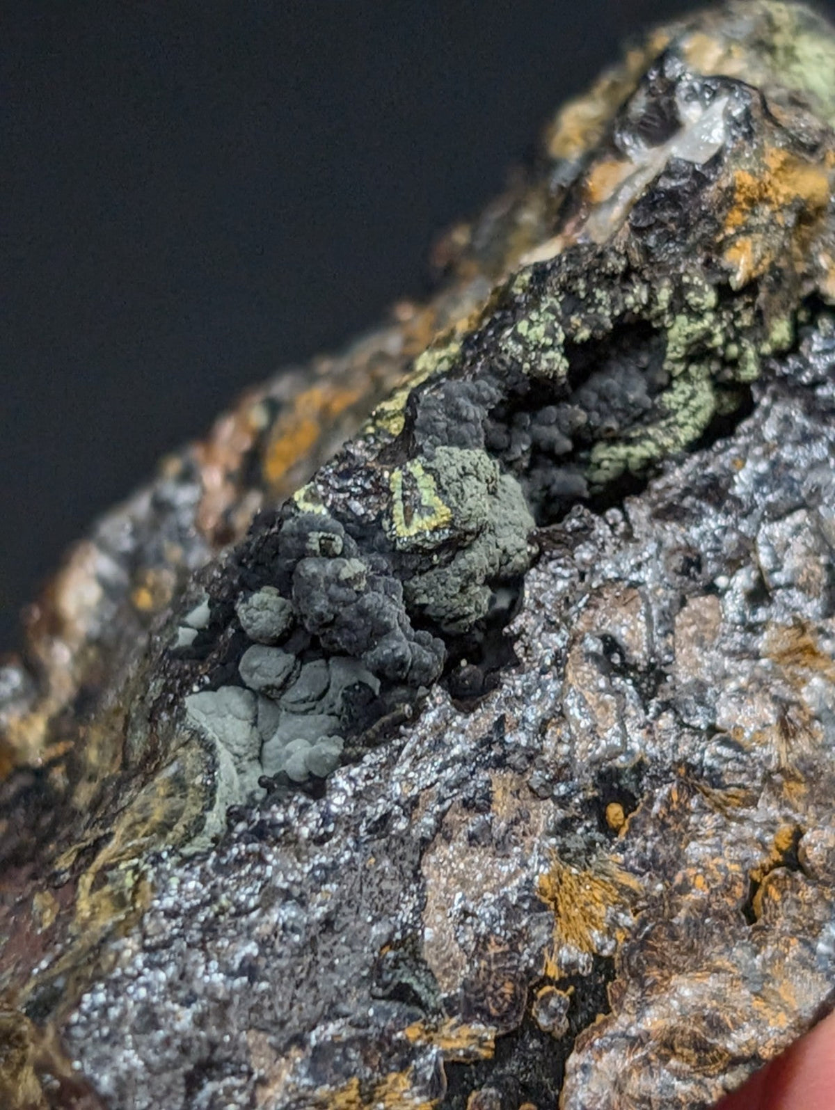 A rugged, textured rock specimen featuring patches of greenish lichen and speckles of orange mineral deposits against a dark, mottled surface.