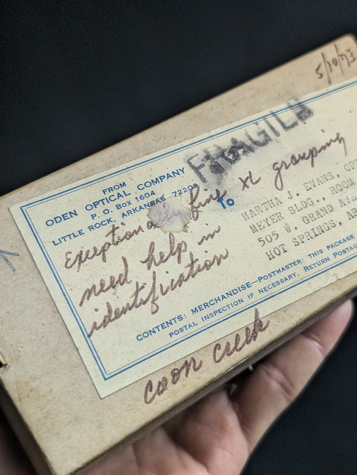 A weathered beige cardboard box with a faded blue-bordered label from oden optical company, featuring handwritten notes and postage markings.