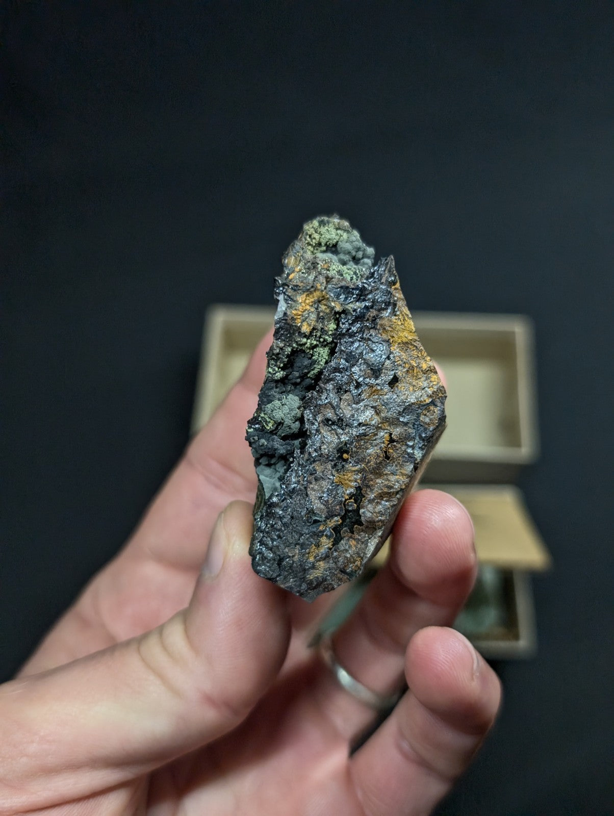 A hand holds a rough, irregularly shaped mineral specimen with dark gray and black surfaces accented by patches of greenish-blue and yellow-orange patina.