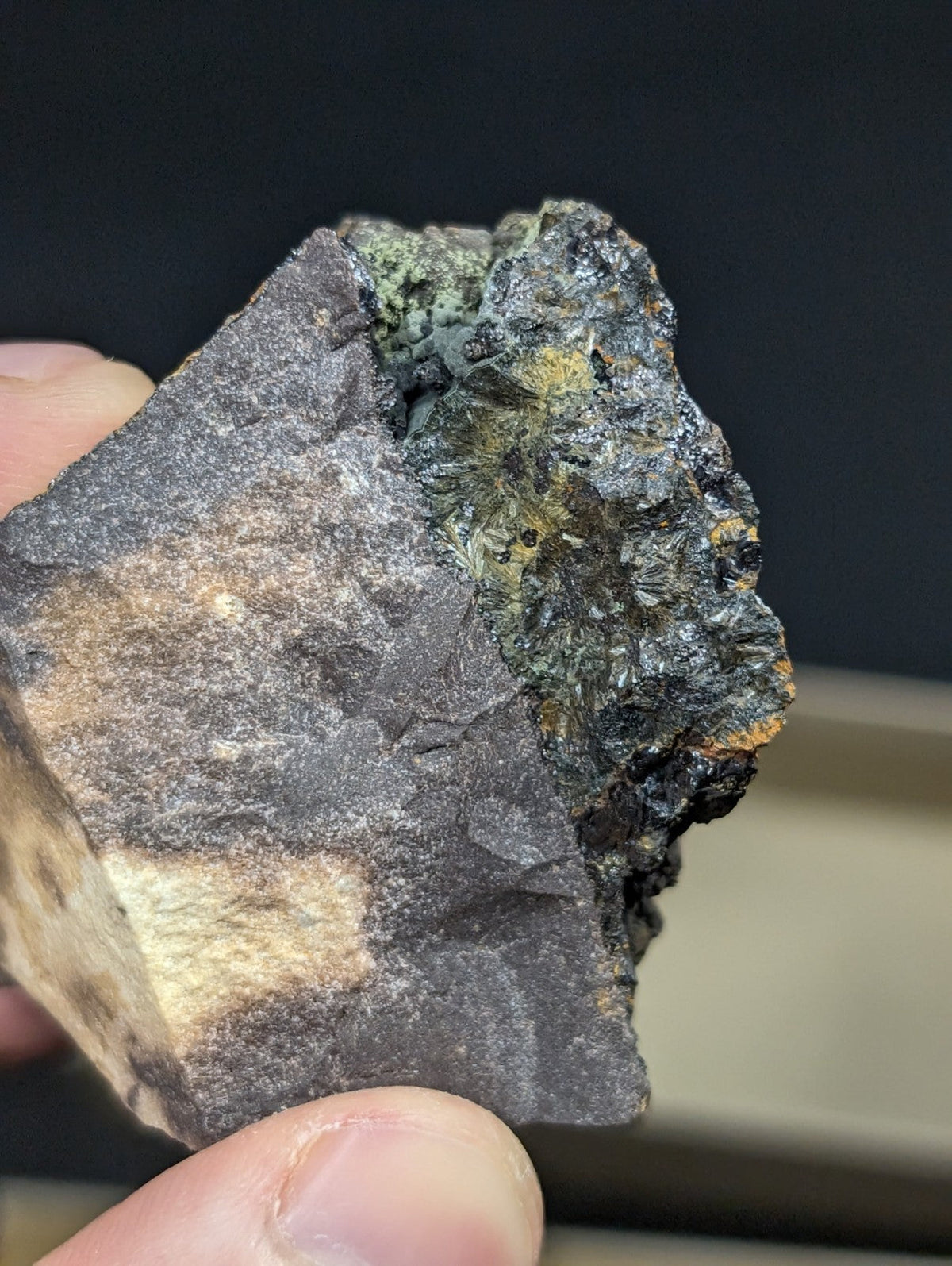 A rugged, dark gray rock fragment with a rough texture and patches of greenish-yellow mineral growth.