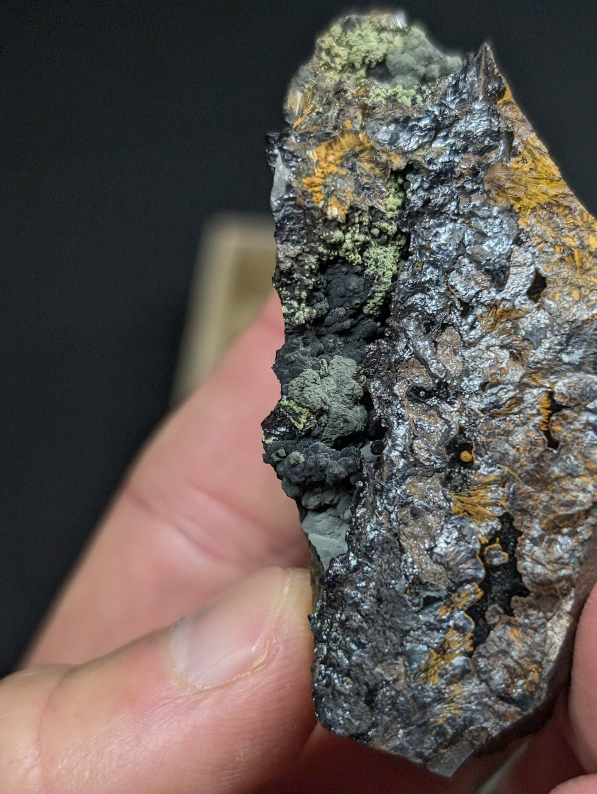 A rugged, irregularly shaped mineral specimen with a dark metallic surface accented by patches of green and orange lichen.