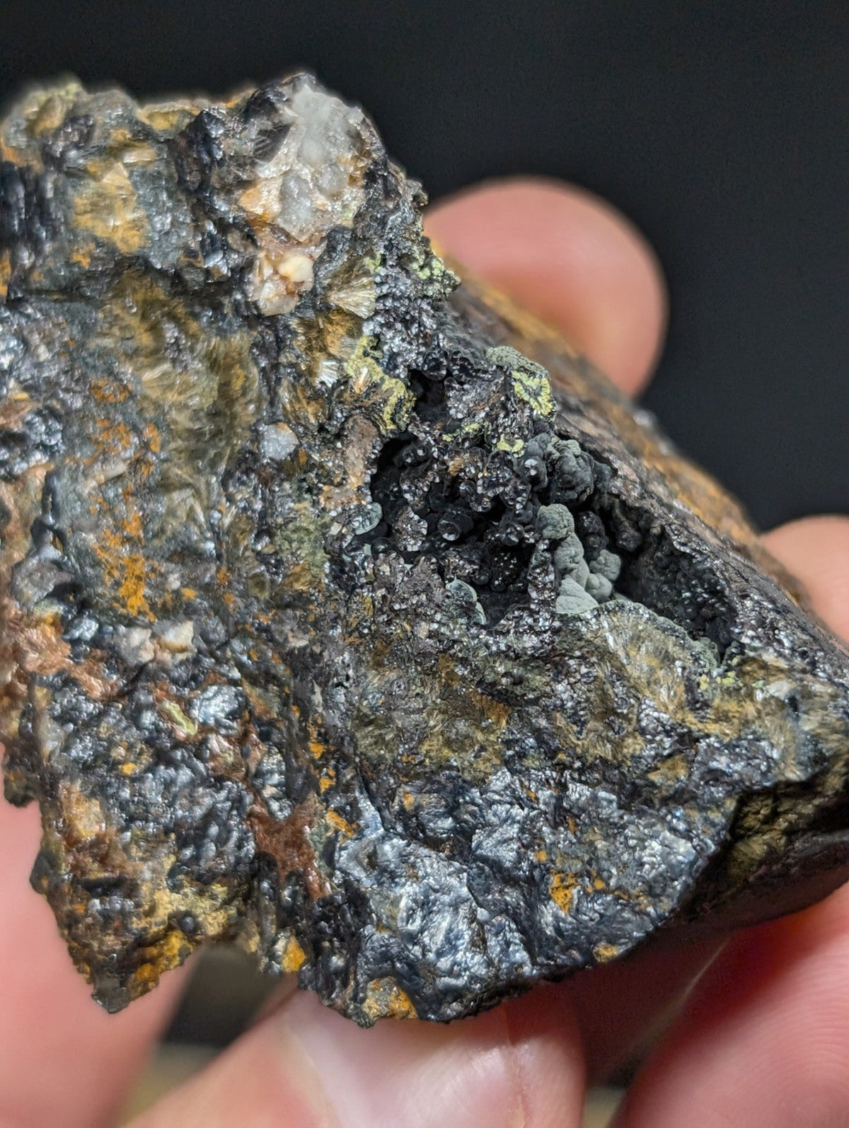 A rugged, irregularly shaped mineral specimen with a dark gray and black surface accented by patches of greenish lichen-like growths and subtle orange-yellow streaks.