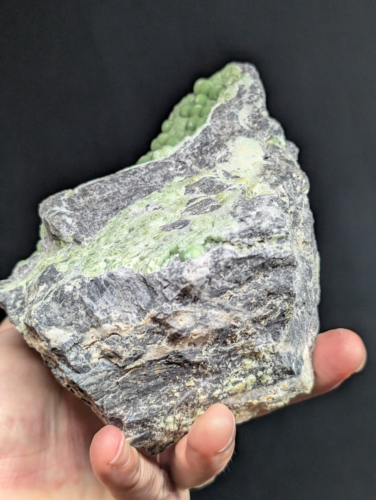 A hand holds a rough, irregularly shaped mineral specimen featuring patches of vibrant green veining against a dark gray and white matrix.