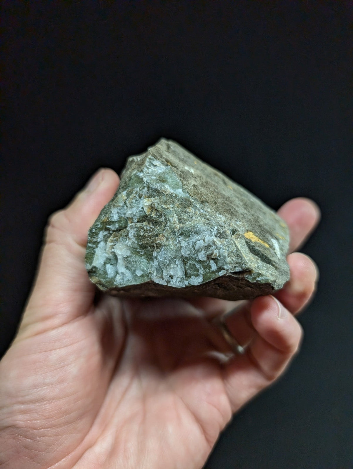 A rough, irregularly shaped greenish-gray rock with subtle white veining and a small patch of yellow mineral inlay.