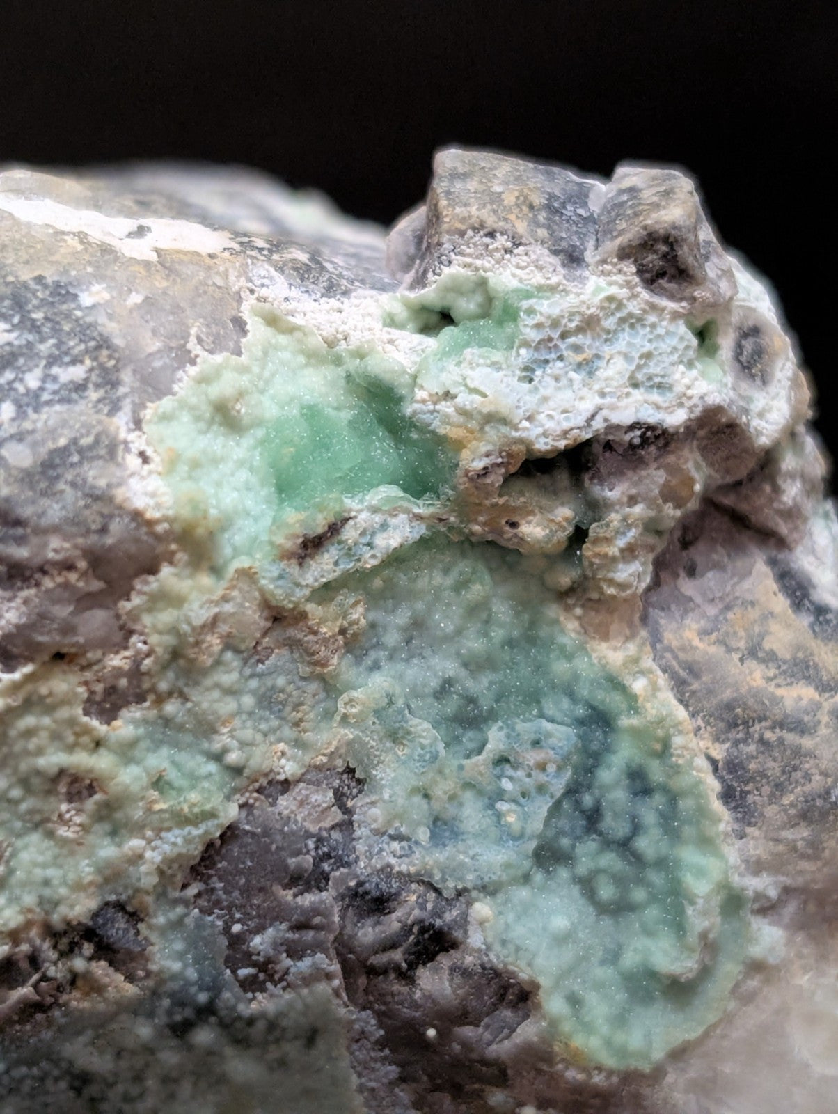 A rough, naturally occurring mineral specimen featuring patches of translucent light green jade-like material embedded in a matrix of gray and beige rock.