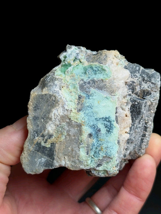 A hand holds a rough, irregularly shaped mineral specimen featuring patches of vibrant turquoise and green hues over a darker grayish base with white crystalline textures.