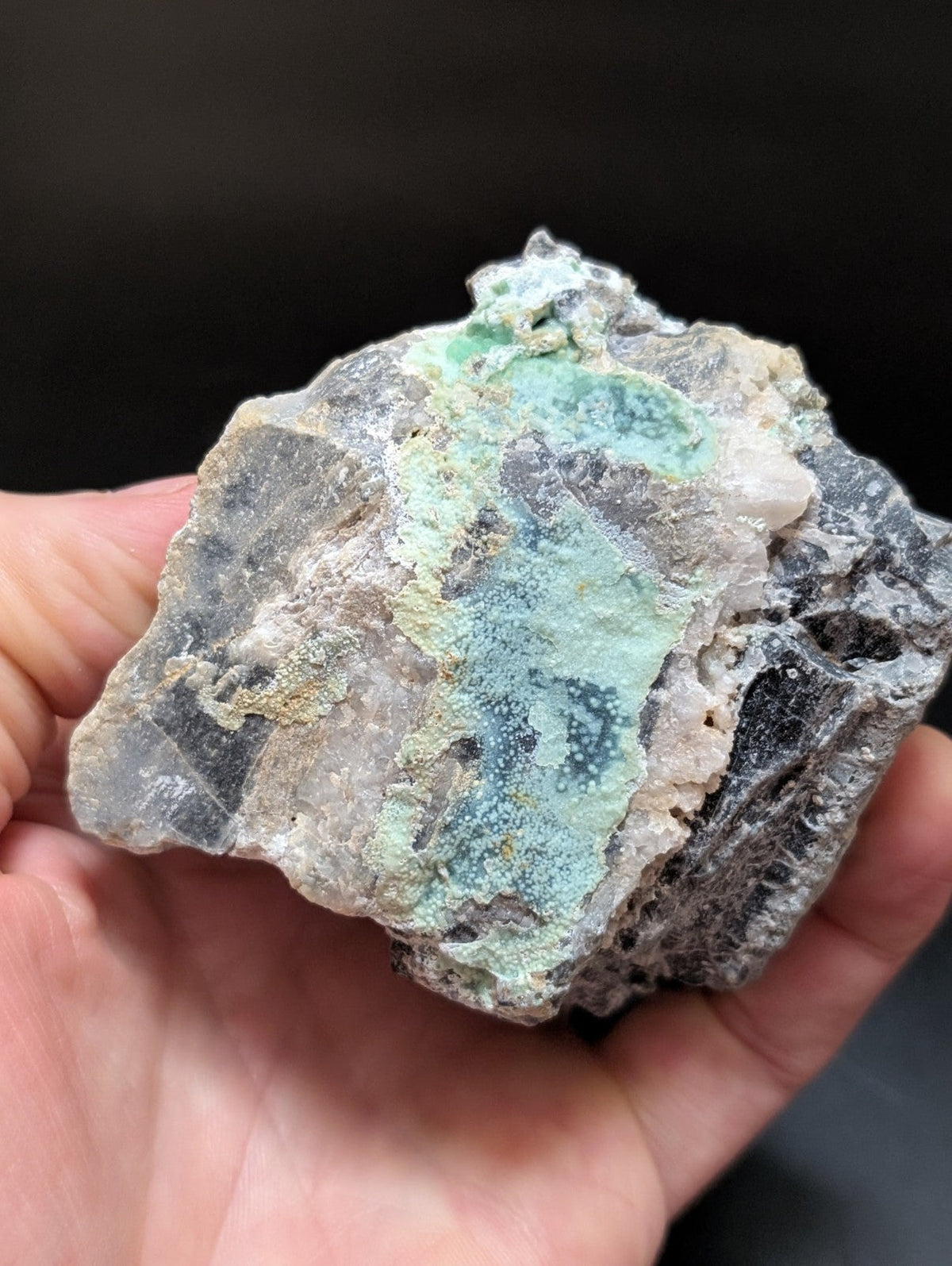 A hand holds a rough, irregularly shaped mineral specimen featuring patches of vibrant turquoise-green veining against a dark gray and black matrix.