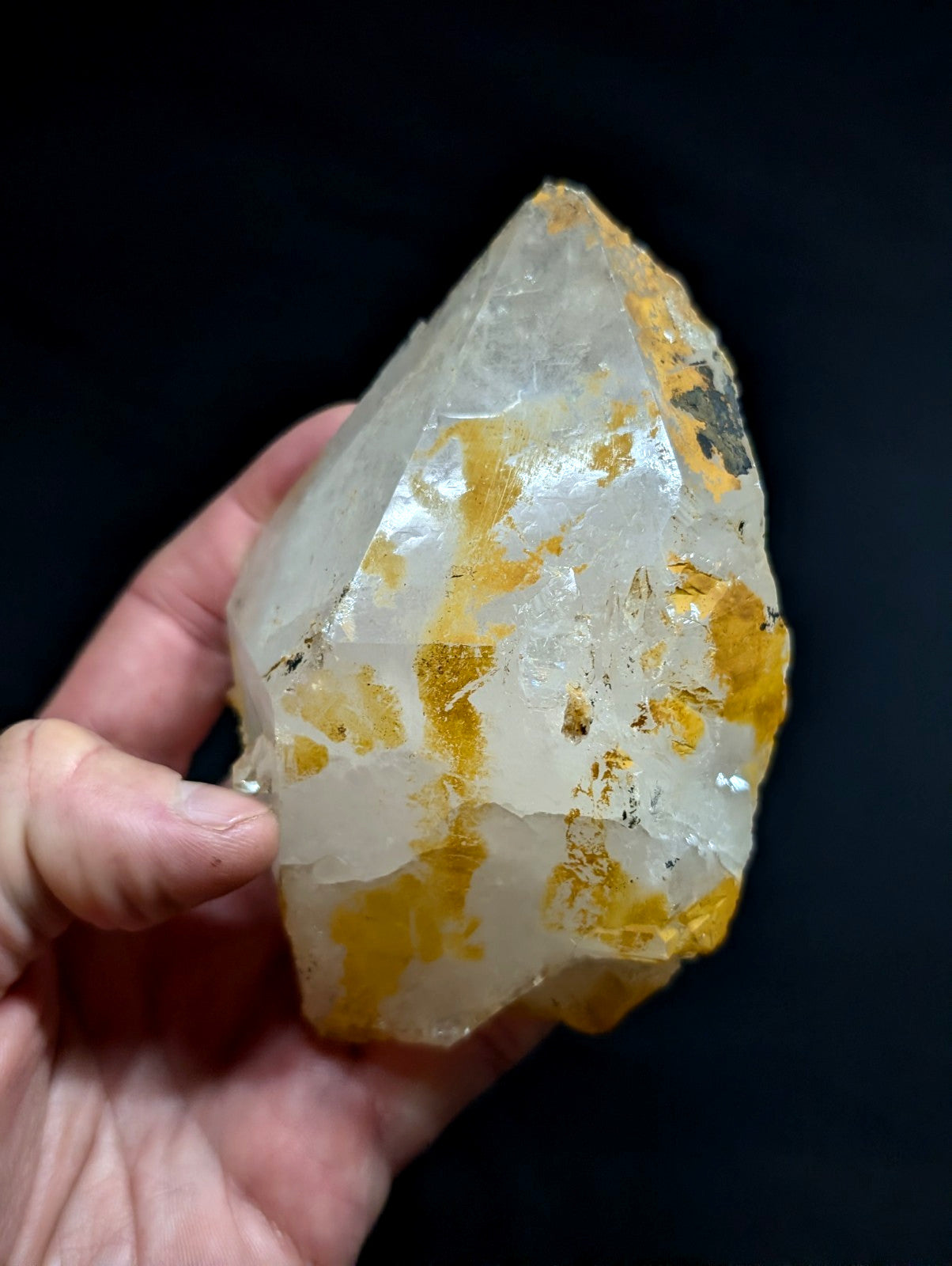 A translucent quartz crystal with golden-yellow inclusions, held gently in a hand against a dark background.