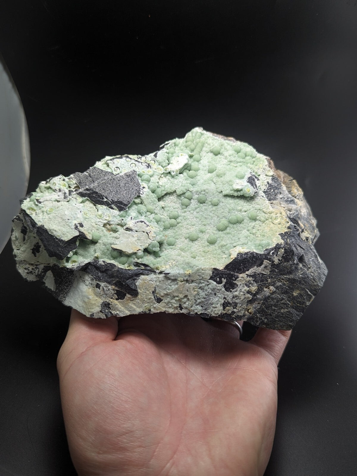A hand holds a rough, irregularly shaped mineral specimen featuring a striking greenish surface with rounded nodules and patches of dark gray rock.