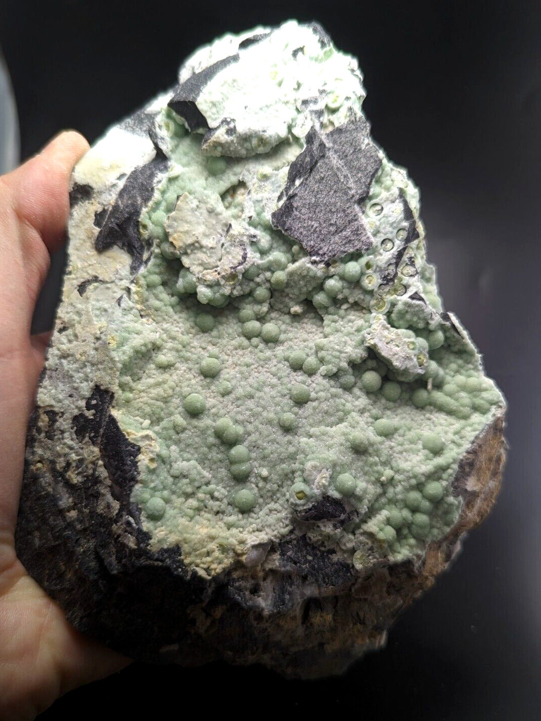 A rough, irregularly shaped mineral specimen featuring a textured surface with clusters of small greenish spheres and patches of grayish-black rock.