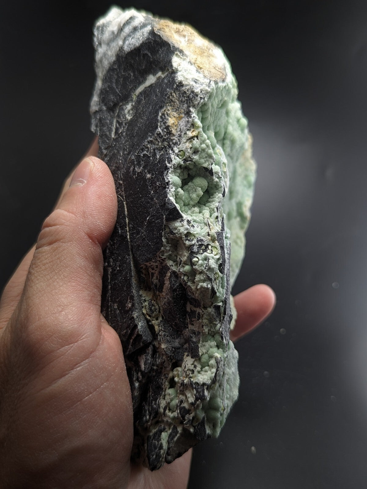 A hand holds a rugged, dark gray rock specimen with vibrant green mineral crystals growing in dense clusters along its surface.