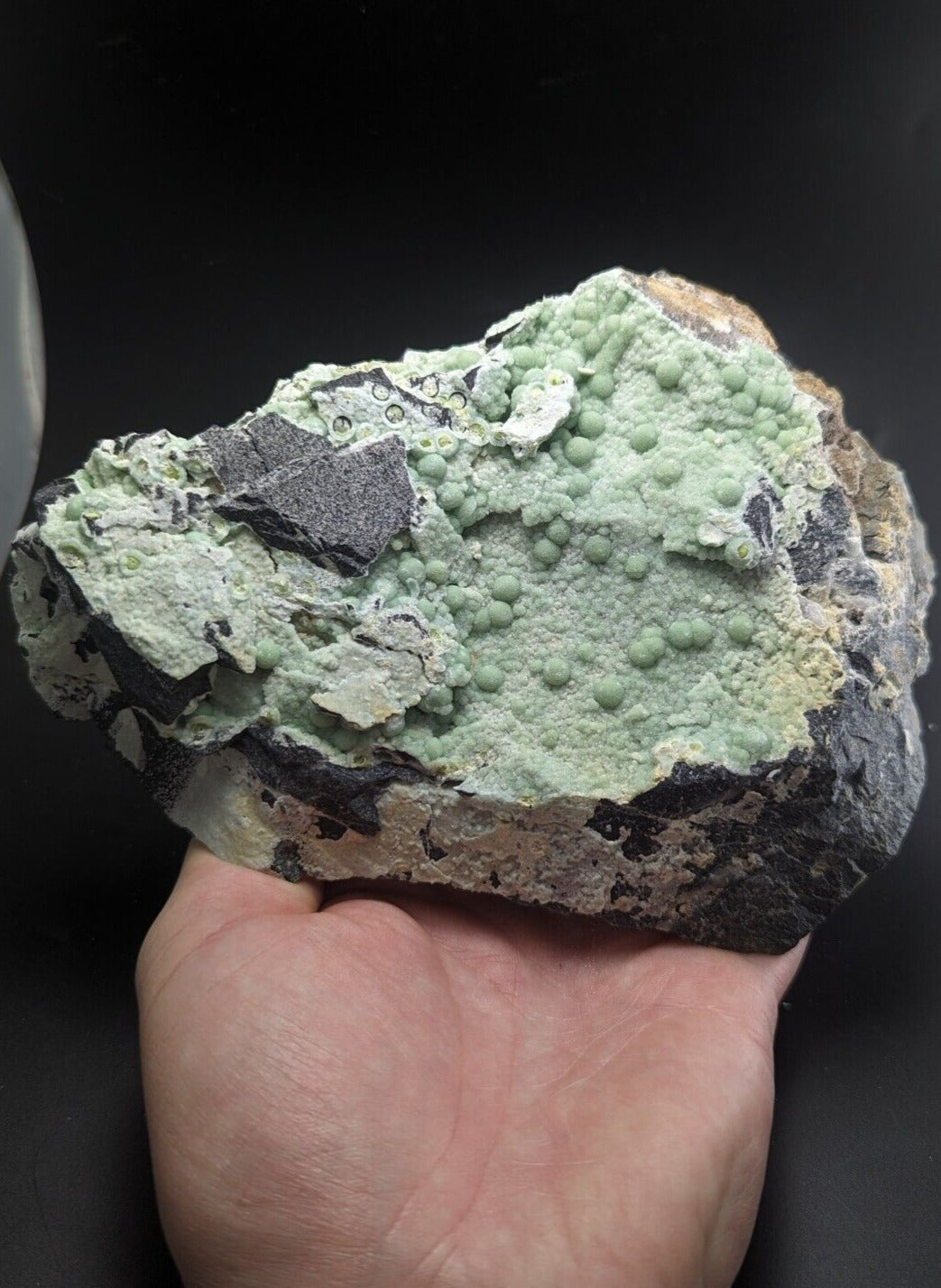 A hand holds a rough, irregularly shaped mineral specimen featuring a prominent light green, bumpy surface with small rounded nodules and patches of darker gray rock.