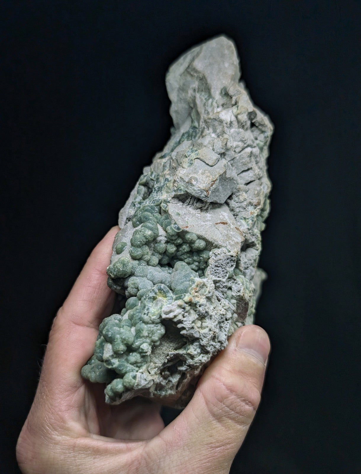 A hand holds a rough, irregularly shaped mineral specimen featuring clusters of greenish-blue nodules against a lighter gray matrix.