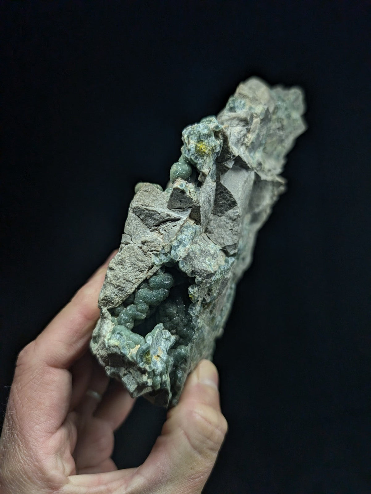 A hand holds a rough, grayish-green mineral specimen featuring clusters of small, rounded green crystals embedded in its rocky matrix.