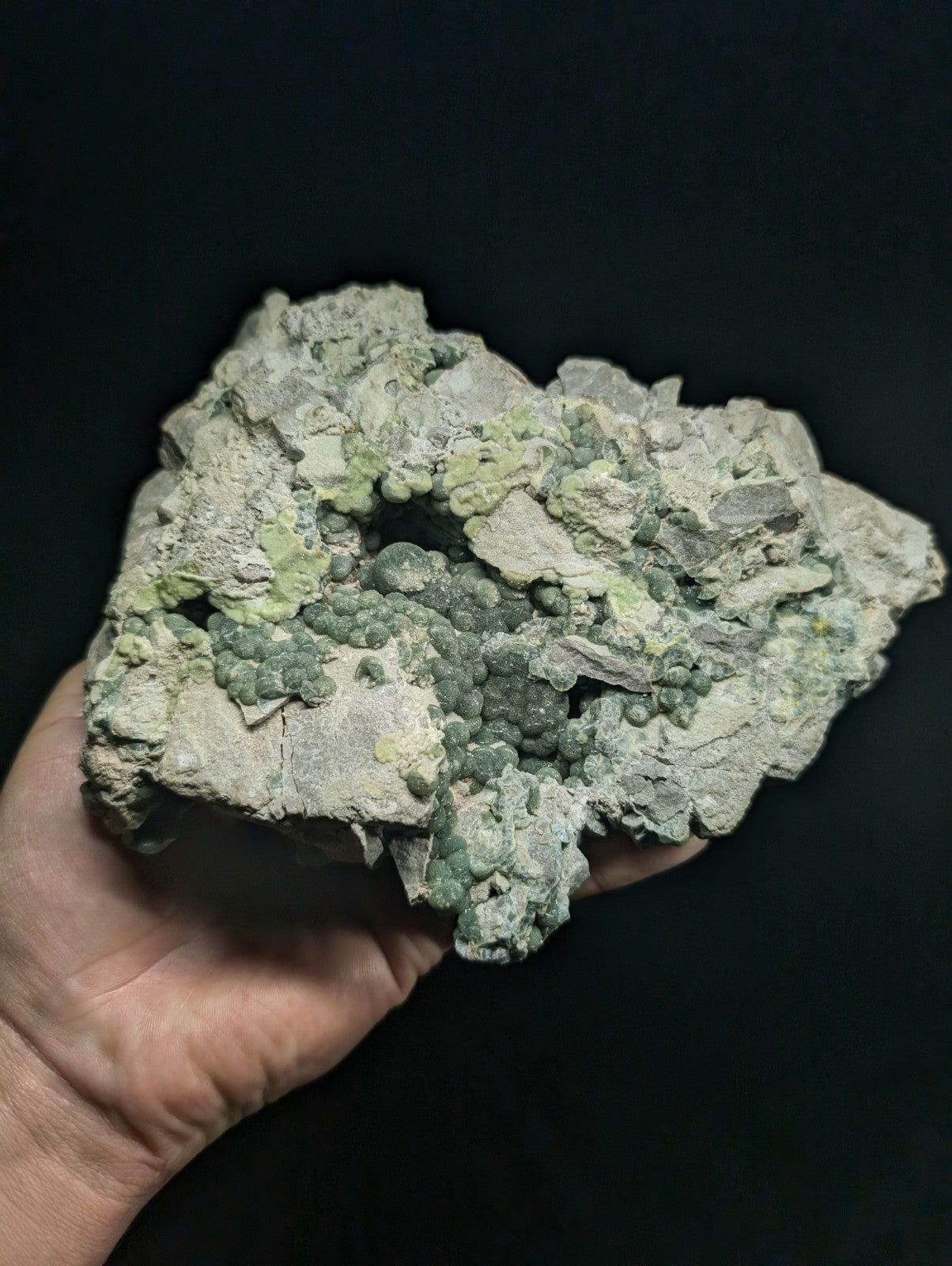 A hand holds a rough, irregularly shaped mineral specimen featuring clusters of greenish-blue crystals growing from a pale gray matrix.