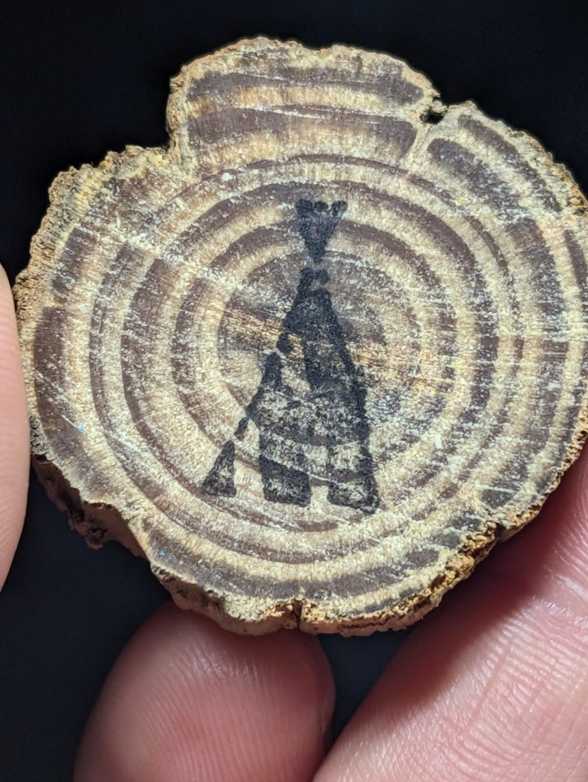 A rustic, circular wooden slice with natural grain patterns and a dark inked silhouette of a figure holding an object.