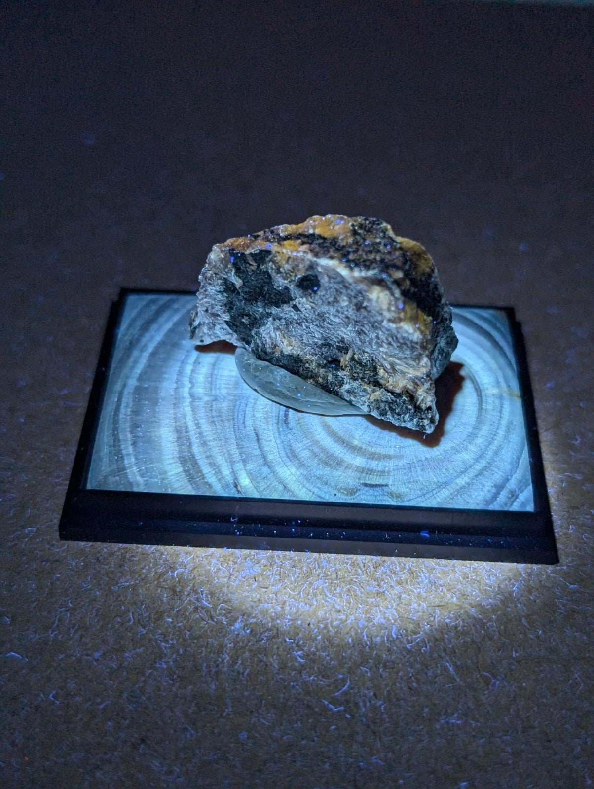 A rugged, irregularly shaped mineral specimen with a dark gray base and patches of yellowish-orange oxidation resting on a circular wooden display stand.