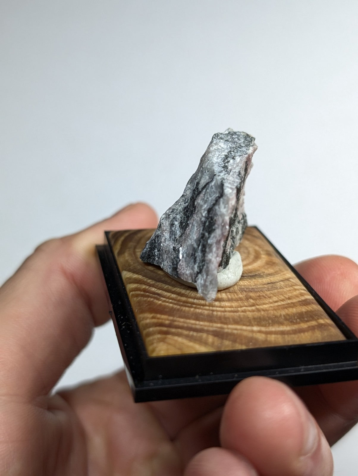 A gray, textured mineral specimen with subtle reddish undertones is displayed on a wooden base within a black frame.