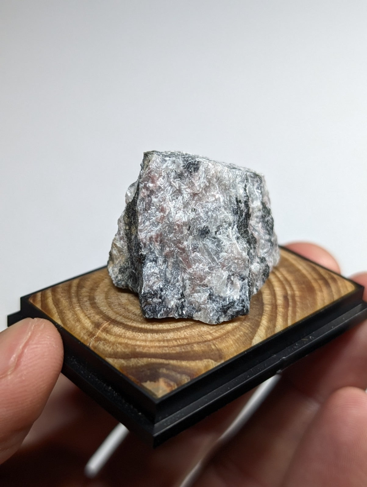 A rough, irregularly shaped gray and black mineral specimen with a speckled surface rests on a square wooden display base.