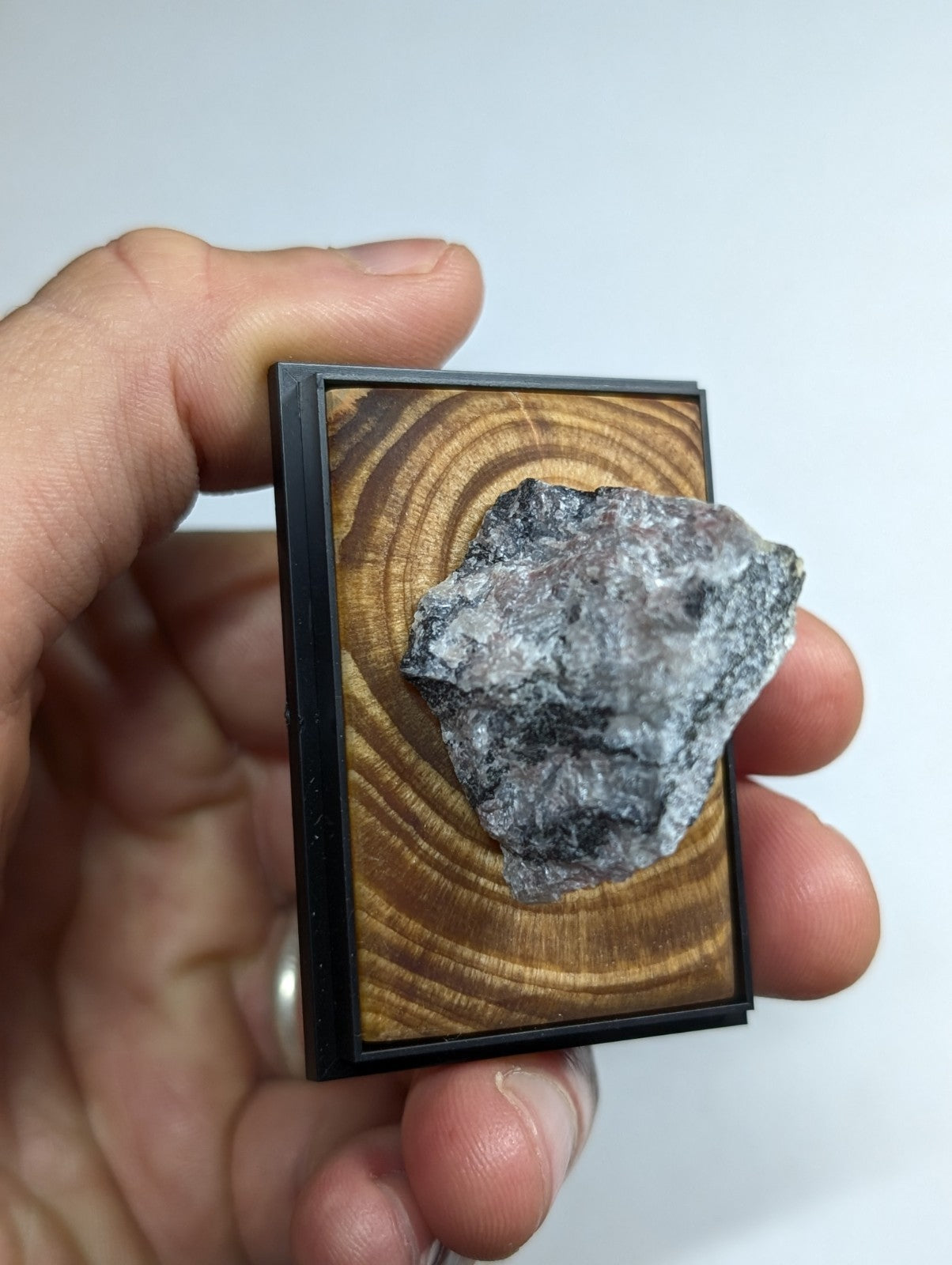 A rough, grayish-black mineral specimen displayed in a black-framed wooden display case with visible grain.