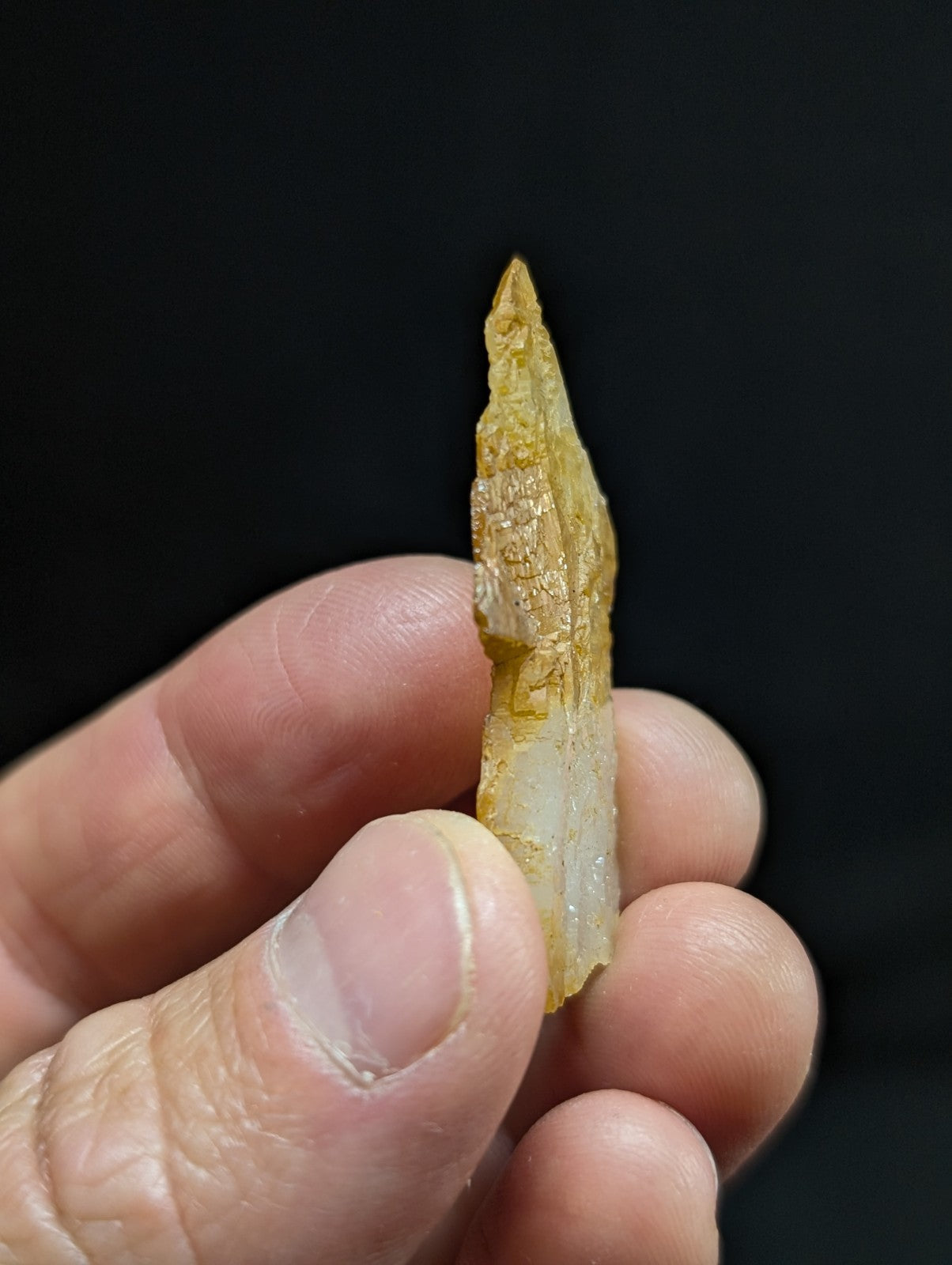 A translucent yellowish-white jadeite point with a pointed tip and natural surface texture, held delicately between fingers.