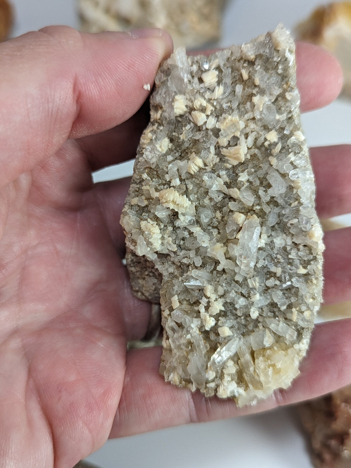 A hand holds a rough, irregularly shaped mineral specimen featuring translucent white and pale yellow crystals embedded in a darker matrix.